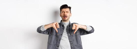 Sad man crying and showing thumbs down, whining and complainin, being upset and disappointed, standing on white background photo