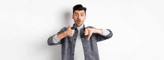 Excited guy show thumbs up and down judging and making decision, rate something interesting, standing on white background photo