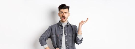 Confused and annoyed guy raising hand up and complaining, asking wtf happened, cant understand, standing on white background photo