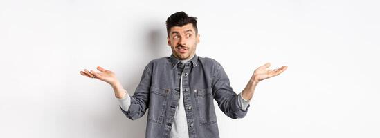 Indecisive young man looking aside and shrugging, know nothing, have no idea, standing clueless and confused against white background photo