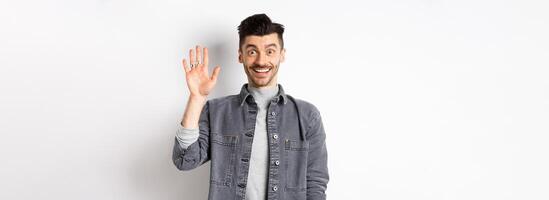 Friendly stylish man saying hello and waiving hand, smiling cheerful, greeting you with hi gesture, standing on white background photo