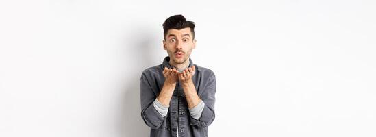 Romantic handsome man blowing air kiss, looking silly at camera, standing in trendy clothes on white background photo