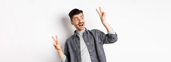 Funny young man showing v-signs and smiling at left side, looking at empty space with cheerful face expression, standing on white background photo
