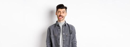 Funny guy with moustache showing tongue and winking, making you laugh, standing on white background photo