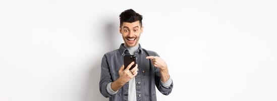 Excited handsome man showing cool news on phone, pointing at smartphone and smiling happy, standing against white background photo