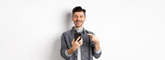Happy handsome caucasian man with moustache pointing finger at smartphone, smiling and showing good deal online on mobile phone, standing pleased against white background photo