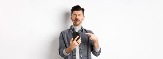 Sad crying guy pointing at smartphone and sobbing, complaining or feeling miserable, standing on white background photo