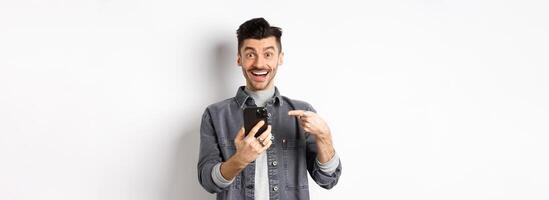 Excited young man receive good news on phone, pointing at smartphone and smiling happy, standing on white background photo
