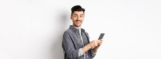 Handsome young man using smartphone and looking at camera. Caucasian guy chatting on phone with happy face, standing on white background photo