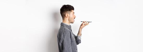 Profile of young man speaking at smartphone, translate with voice-translator app, talking into mobile phone speakerphone, standing on white background photo