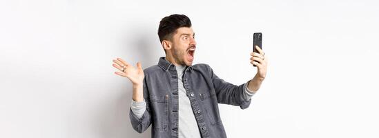 Shocked and confused man staring frustrated at smartphone screen, shouting from disappointment, standing against white background photo