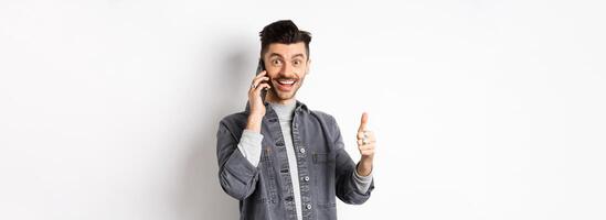 Excited young man talking on phone and showing thumbs up, smiling satisfied, standing against white background photo