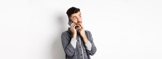 Image of pensive handsome man talking on phone and thinking, making choice while making takeout order, standing against white background photo