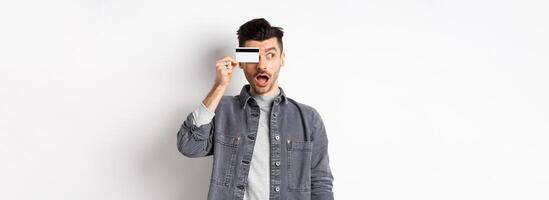 Excited funny guy look aside at logo and show plastic credit card, standing amazed on white background photo