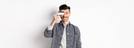 Happy handsome man with moustache show plastic credit card on eye, smiling excited at camera, standing on white background photo