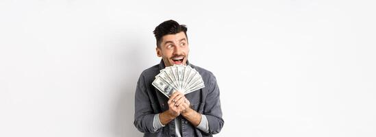 Rich happy man dreaming of shopping wish dollar bills in hands, looking aside pensive and holding money, standing against white background photo