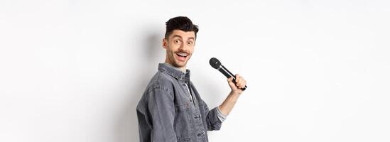 Profile of handsome smiling man holding mic, turn head at camera with excited face, singing karaoke and perform standup, standing on white background photo