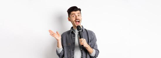 Excited stylish guy giving speech, talking in microphone, perform or sing karaoke, standing on white background photo