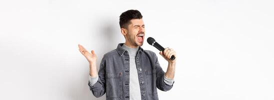 Handsome guy singing in mic with passion, perform for audience, holding microphone and gesturing, standing on white background photo