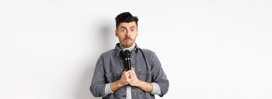 Nervous unconfident guy holding microphone and look indecisive, scared to perform, standing shy against white background photo