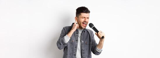 Handsome singer trying to reach high note, singing in microphone and shut one ear, standing on white background photo