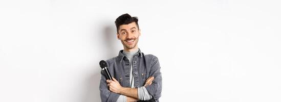 Handsome caucasian guy with moustache cross arms on chest, holding mic and smiling at camera, perform on stage with microphone, standing against white background photo