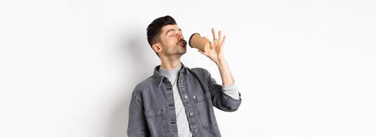 Funny guy enjoying drinking coffee from cup, sipping takeaway drink from cafe with satisfaction, standing on white background photo