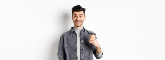 Enthusiastic handsome man holding coffee cup and smiling, drinking good drink from cafe takeout, standing against white background photo