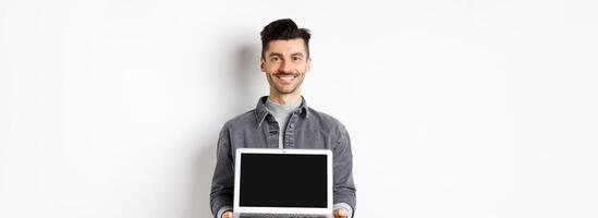 Smiling young man showing empty laptop screen, showing online deal, standing on white background photo