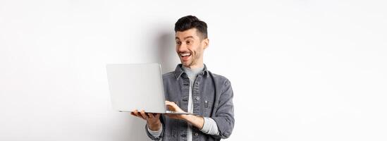 Man watching funny on laptop, laughing at computer screen and smiling joyful, standing on white background photo