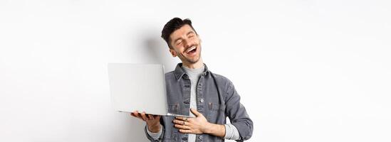 Image of happy guy laughing at funny on laptop, touching belly and smiling satisfied wth good laugh, standing against white background photo