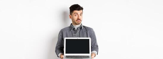 Indecisive young man pouting and shrugging while showing empty laptop screen, standing clueless on white background photo