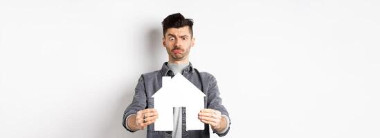 Real estate and insurance concept. Confused guy look at paper house cutout unsure, frowning perplexed, standing on white background photo