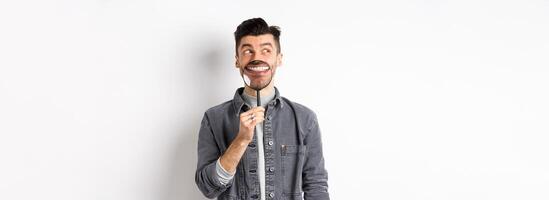 Handsome positive guy showing white perfect smile with magnifying glass, looking left at logo, standing against white background photo