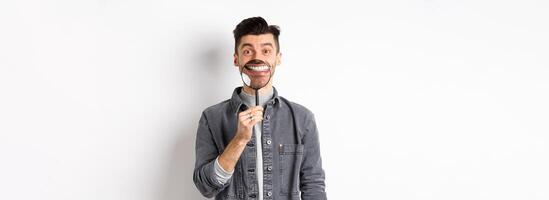 Handsome positive guy showing white perfect smile with magnifying glass, standing against white background photo