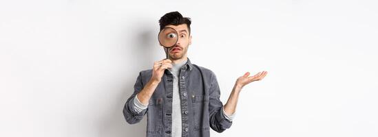 Confused man look through magnifying glass and shrugging clueless, standing on white background photo