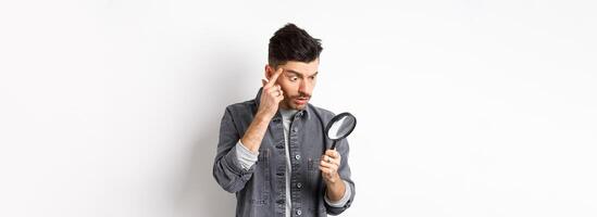 Man looking through magnifying glass and stretching eyelid to see clearly, standing on white background photo