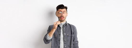 Funny young man squinting, showing faces with magnifying glass, standing on white background photo