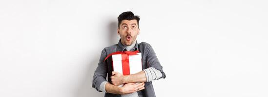 Surprised funny guy hugging big holiday gift and looking up, say wow amazed, checking out Valentines day special offer, standing on white background photo