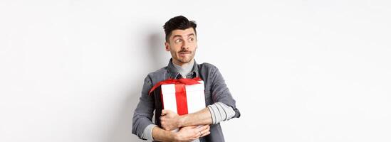 Excited man hugging romantic gift for girlfriend, waiting for valentines date with lover, looking at empty space logo with happy face, standing on white background photo