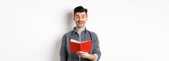 Hobbies and leisure. Happy young man reading planner, holding diary or red journal and smiling, making notes, standing on white background photo
