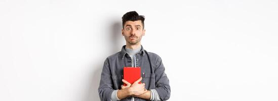 Cute and shy guy hugging his diary, holding red journal and look modest at camera, standing against white background photo