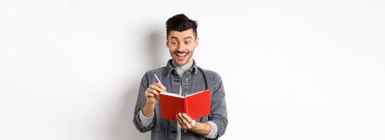 Excited guy writing in planner and smiling, writing down ideas in journal or diary, standing on white background photo