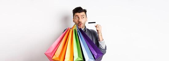 Excited young man buying in shop with plastic credit card, holding colorful shopping bags and looking aside at logo, standing against white background photo