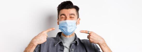 Covid-19, health and lifestyle concept. Cheerful smiling man pointing at sterile medical mask, protect himself from corona outbreak, standing on white background photo