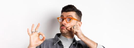 Funny young man in glasses touching his french moustache and showing okay, not bad sign, approve and like good product, standing on white background photo