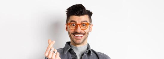 Close up portrait of cute guy with moustache wearing glasses, showing hand heart sign, smiling happy, standing on white background photo