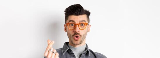 Close up portrait of cute guy with moustache wearing glasses, showing hand heart sign, look impressed at camera, standing on white background photo