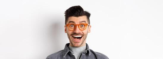 Excited funny man in glasses smiling, look with amazement and joy at awesome promo, standing happy against white background photo