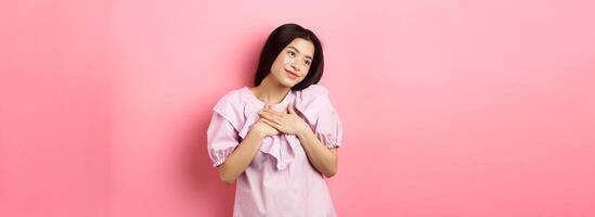 Dreamy asian girl smiling and holding hands on chest, looking left at something romantic and cute, standing on pink background photo